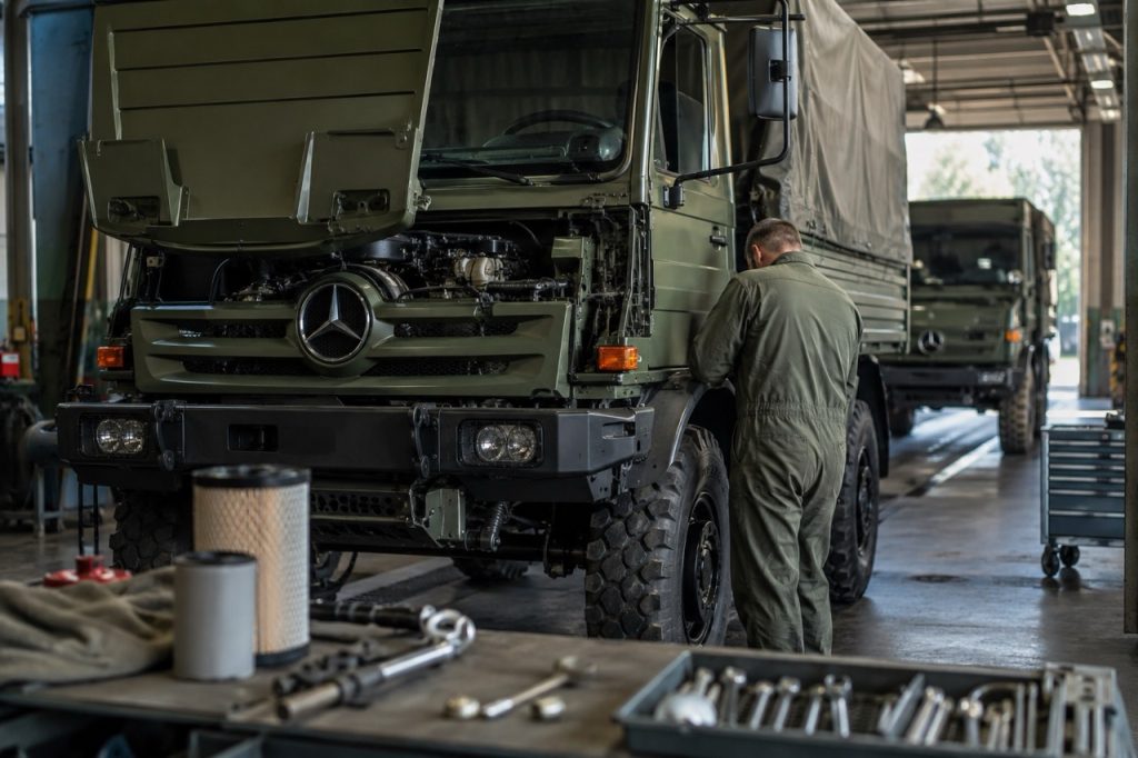Askeri bakım atölyesinde motor kapağı açık Mercedes-Benz Unimog taktik aracına önleyici bakım yapan teknisyen. Düzenli bakım, operasyonel hazırlığı ve araç güvenilirliğini artırır.